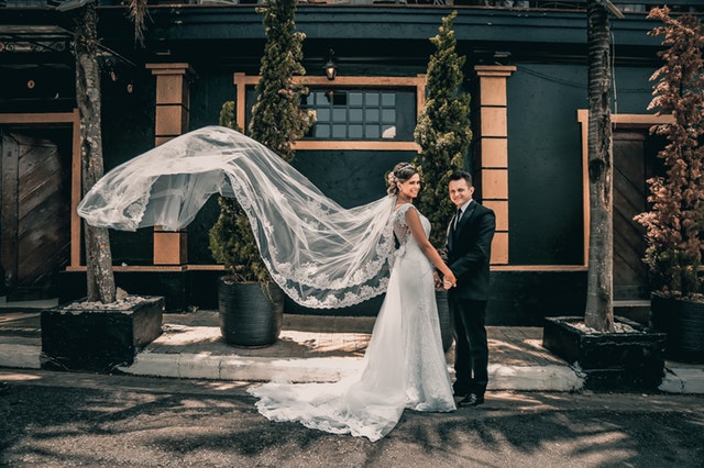Ženích v obleku a nevesta s dlhým závojom stoja pred sivou budovou.jpg